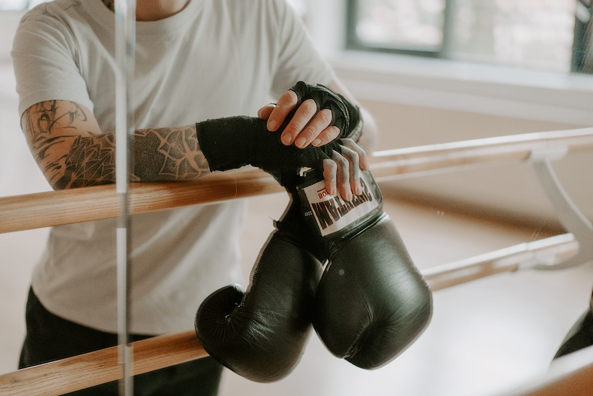 Eine Person mit Boxhandschuhen in der Hand steht in einem Sportstudio an eine Ballettstange gelehnt vor einem Spiegel 