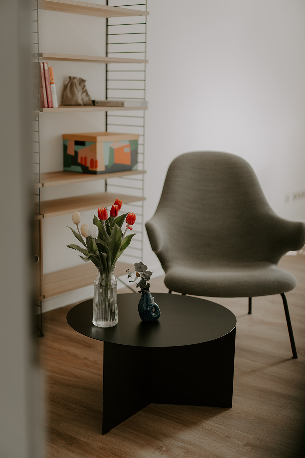 Ein grauer Sessel und ein kleiner schwarzer Beistelltisch mit Blumen darauf vor einem Bücherregal