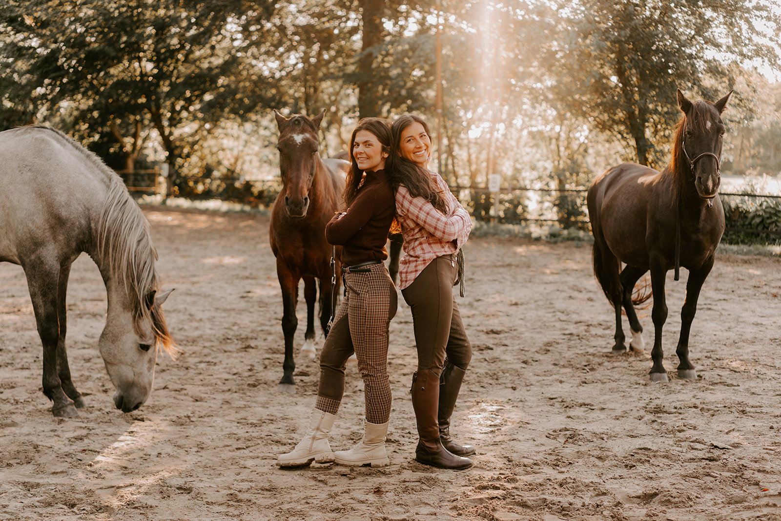 Zwei Familientherapeutinnen stehen Rücken an Rücken auf einem Reitplatz mit zwei Pferden